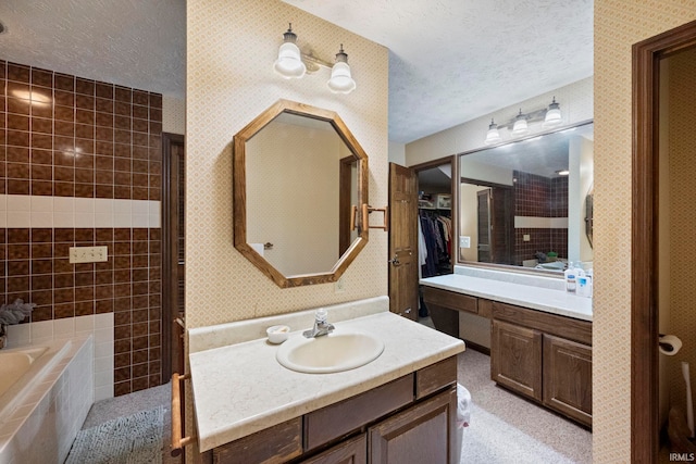 full bathroom featuring a textured ceiling, wallpapered walls, a relaxing tiled tub, two vanities, and a spacious closet