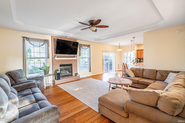 living room with a raised ceiling, ceiling fan, light wood-style floors, a fireplace, and crown molding