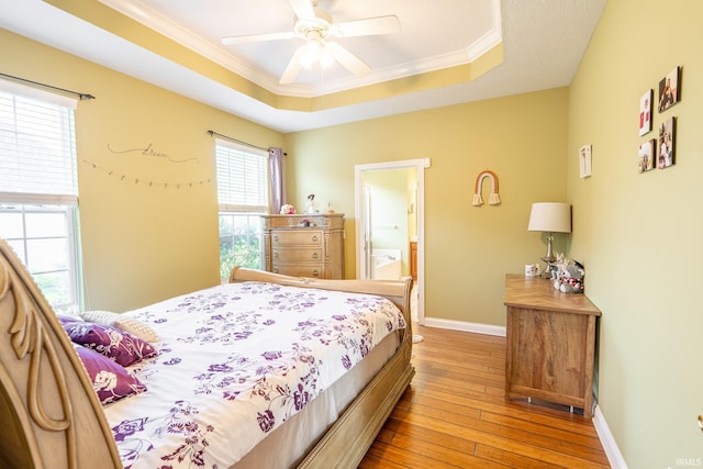 bedroom featuring a tray ceiling, light wood-type flooring, ornamental molding, baseboards, and ensuite bath
