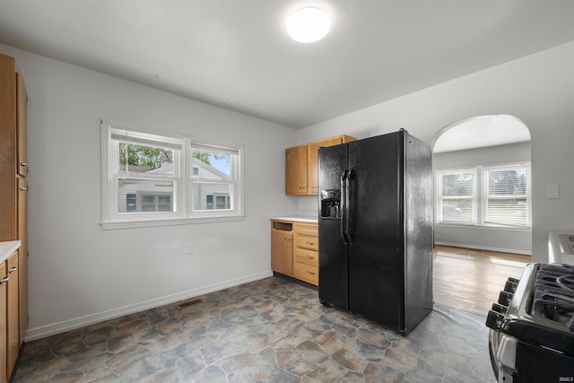 kitchen featuring black refrigerator with ice dispenser, arched walkways, gas range, stone finish flooring, and baseboards