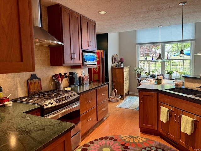 kitchen featuring stainless steel appliances, wall chimney exhaust hood, light wood-style flooring, pendant lighting, and brown cabinetry