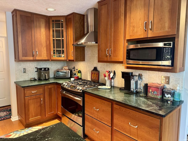 kitchen with wall chimney range hood, stainless steel appliances, a textured ceiling, brown cabinets, and light wood-style flooring