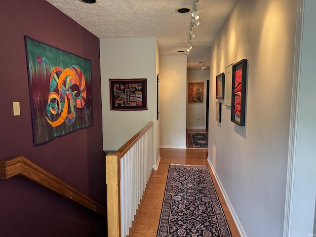corridor featuring wood finished floors, track lighting, a textured ceiling, an upstairs landing, and baseboards