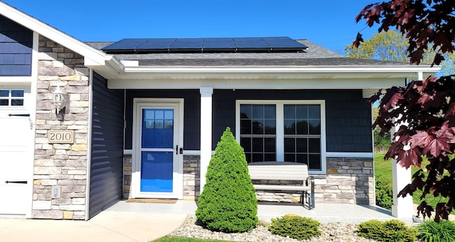view of exterior entry featuring stone siding, covered porch, solar panels, and a shingled roof