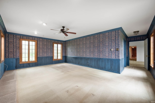 carpeted empty room featuring crown molding, a wainscoted wall, wallpapered walls, and a ceiling fan
