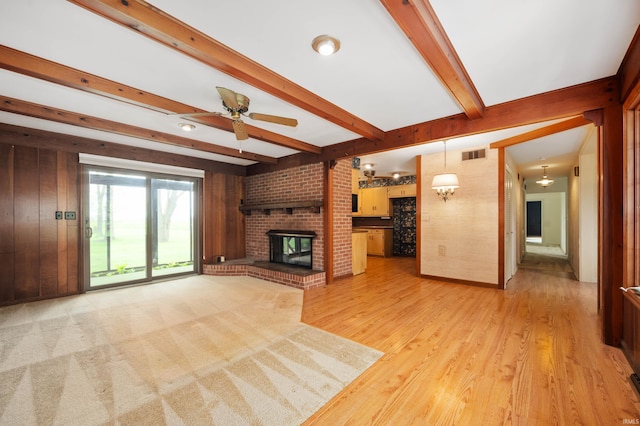 unfurnished living room featuring ceiling fan, beamed ceiling, a brick fireplace, light wood finished floors, and wooden walls