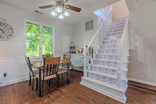 dining room featuring stairs, dark wood-type flooring, ceiling fan, and a wainscoted wall