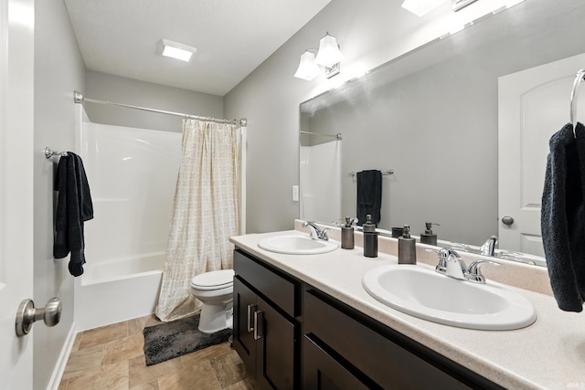 bathroom featuring toilet, shower / bath combination with curtain, stone finish floors, double vanity, and baseboards