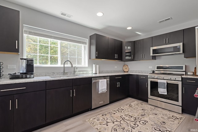 kitchen featuring stainless steel appliances, a sink, glass insert cabinets, dark cabinetry, and light stone counters
