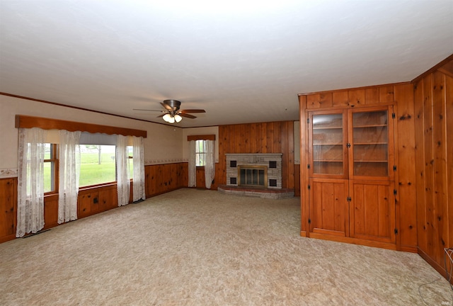 unfurnished living room featuring wood walls, ceiling fan, light carpet, a brick fireplace, and wainscoting