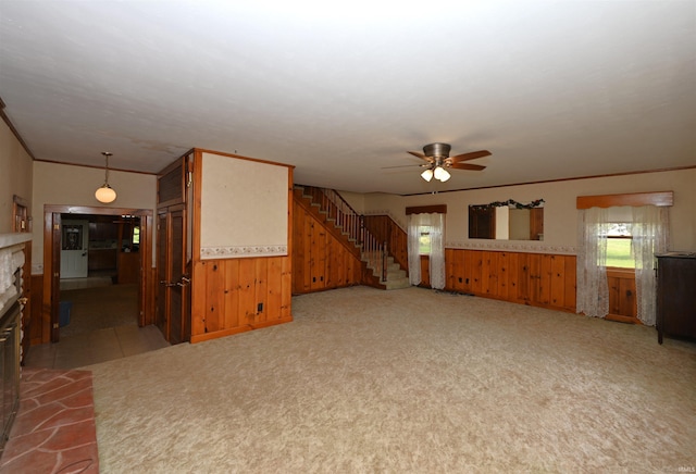 unfurnished living room featuring a ceiling fan, a fireplace, wood walls, stairway, and crown molding