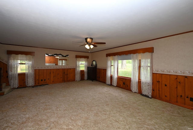 unfurnished living room with wainscoting, ceiling fan, carpet flooring, crown molding, and wooden walls