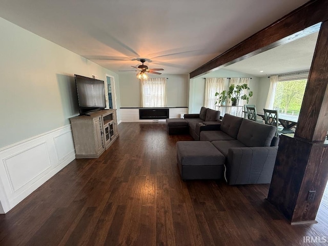 living room with plenty of natural light, dark wood-style floors, a ceiling fan, a wainscoted wall, and beamed ceiling