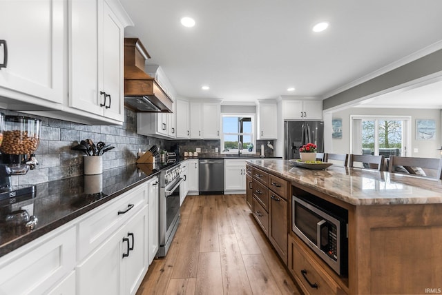 kitchen with appliances with stainless steel finishes, premium range hood, healthy amount of natural light, dark stone countertops, and crown molding