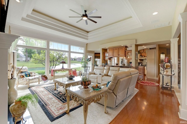 living room with a tray ceiling, a ceiling fan, a water view, light wood finished floors, and ornate columns