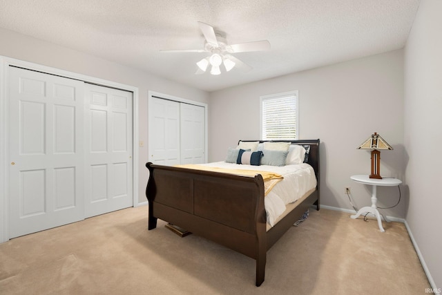bedroom featuring light colored carpet, two closets, a textured ceiling, baseboards, and ceiling fan