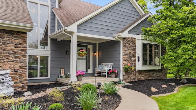 view of exterior entry featuring stone siding, covered porch, and a shingled roof