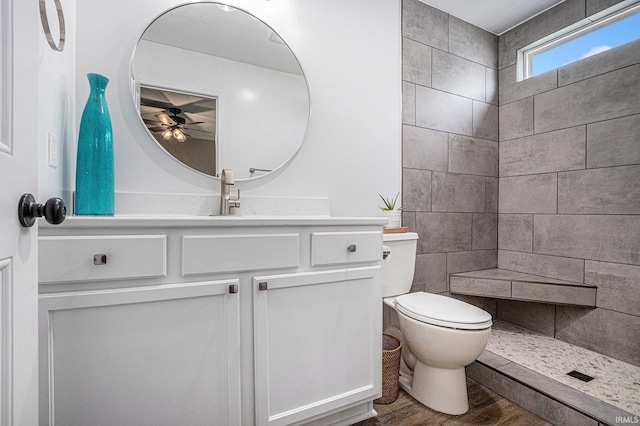 bathroom with toilet, wood finished floors, vanity, a ceiling fan, and a tile shower