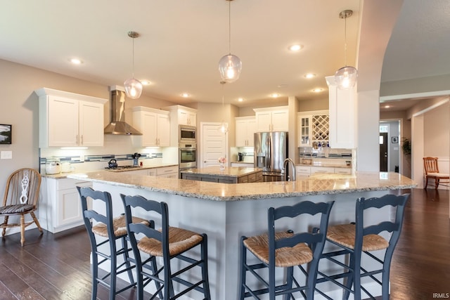kitchen featuring wall chimney exhaust hood, stainless steel appliances, a spacious island, dark wood finished floors, and a breakfast bar area