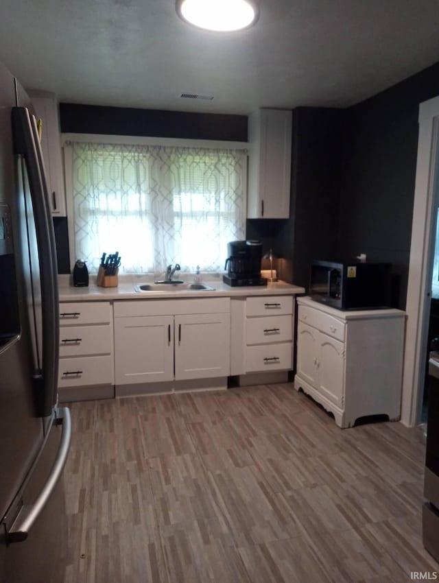 kitchen with stainless steel fridge, black microwave, white cabinetry, a sink, and light wood-style flooring