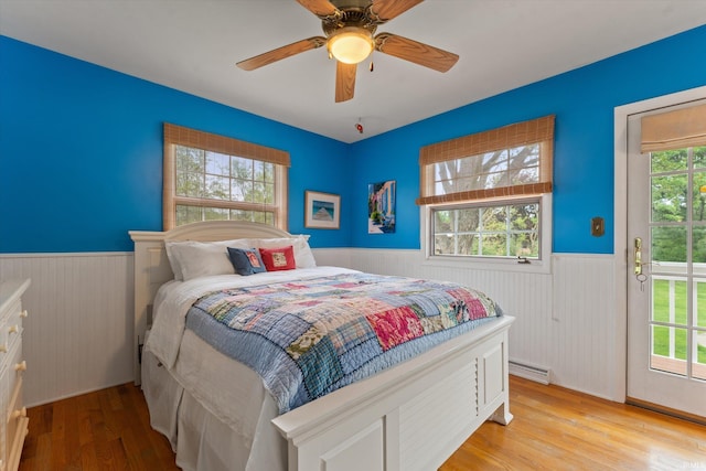 bedroom with a wainscoted wall, access to exterior, light wood-style flooring, a baseboard heating unit, and a ceiling fan