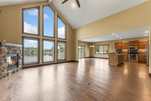 unfurnished living room with a ceiling fan, high vaulted ceiling, a stone fireplace, dark wood-type flooring, and a water view