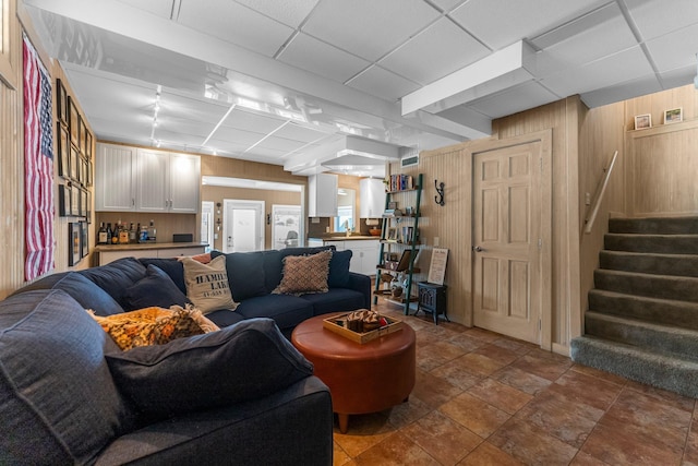 living room featuring stairs, wooden walls, stone finish flooring, and a drop ceiling