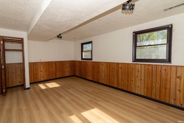 unfurnished room featuring a textured ceiling, a wainscoted wall, light wood-style flooring, healthy amount of natural light, and wooden walls
