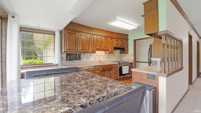 kitchen with stainless steel gas range, under cabinet range hood, brown cabinets, tasteful backsplash, and a sink