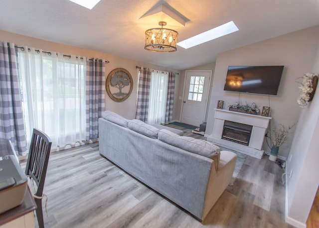 living area featuring a skylight, vaulted ceiling, a chandelier, a glass covered fireplace, and light wood-type flooring