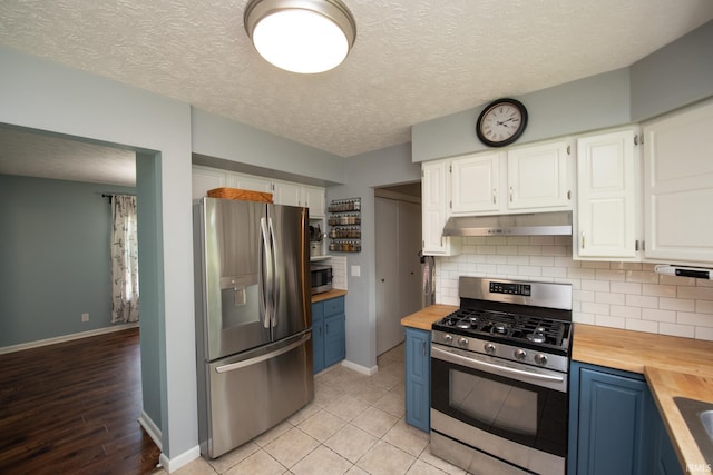 kitchen featuring appliances with stainless steel finishes, butcher block counters, blue cabinetry, under cabinet range hood, and a textured ceiling