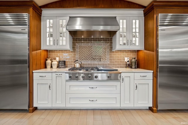 kitchen with appliances with stainless steel finishes, wall chimney range hood, light countertops, glass insert cabinets, and white cabinetry