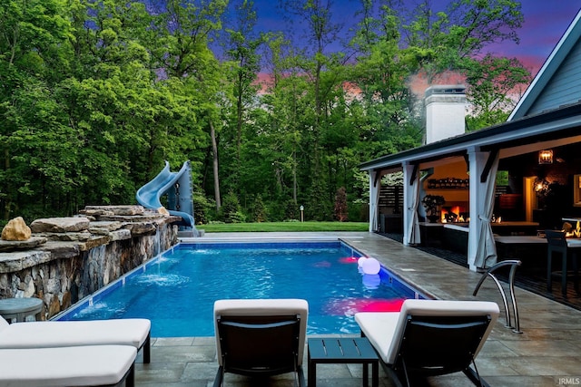 outdoor pool with a water slide, a patio, and a warm lit fireplace