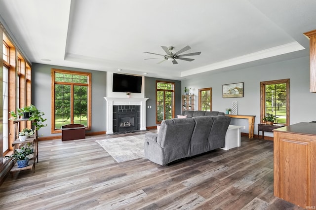 living area featuring a tray ceiling, wood finished floors, ceiling fan, a tiled fireplace, and baseboards