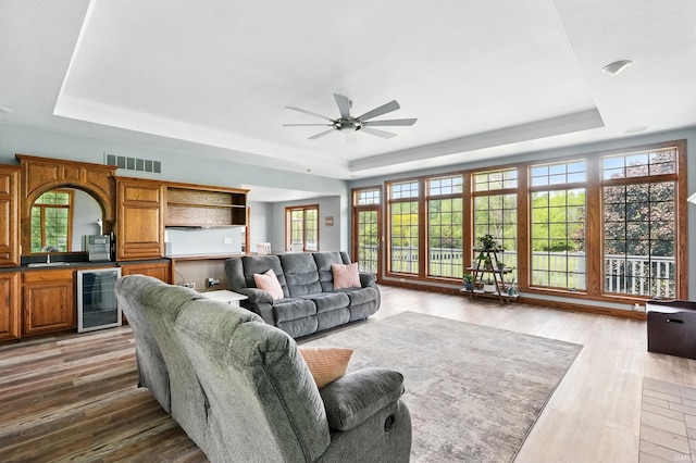 living area with a raised ceiling, wine cooler, a ceiling fan, and wood finished floors