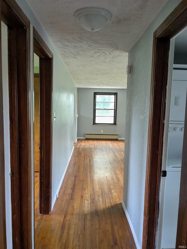 hall featuring stacked washing machine and dryer, a baseboard heating unit, a textured ceiling, wood-type flooring, and baseboards