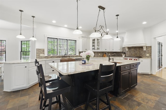 kitchen featuring custom exhaust hood, a sink, stone tile flooring, an island with sink, and recessed lighting