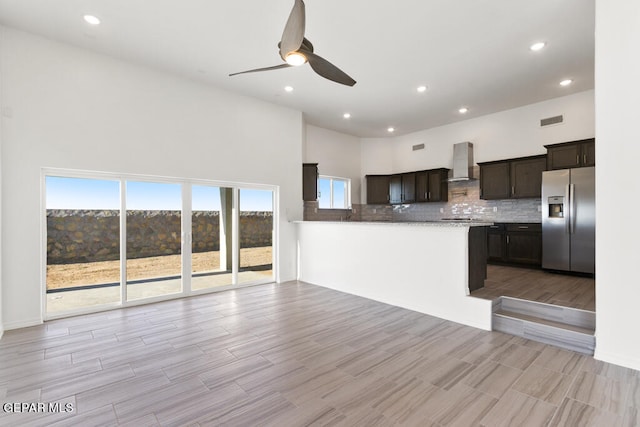 kitchen featuring backsplash, ceiling fan, stainless steel fridge with ice dispenser, a towering ceiling, and wall chimney range hood