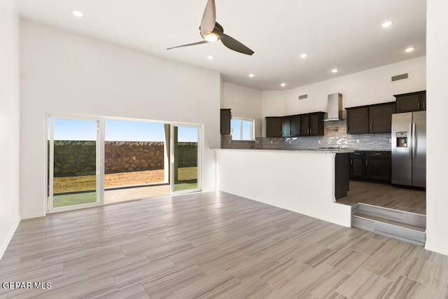 kitchen with backsplash, ceiling fan, stainless steel fridge, light stone counters, and wall chimney exhaust hood