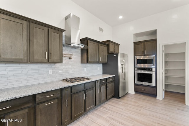 kitchen featuring appliances with stainless steel finishes, light stone countertops, wall chimney exhaust hood, tasteful backsplash, and dark brown cabinetry