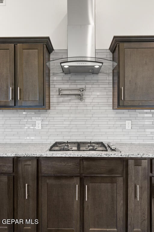 kitchen featuring stainless steel gas stovetop, tasteful backsplash, dark brown cabinets, wall chimney exhaust hood, and light stone counters