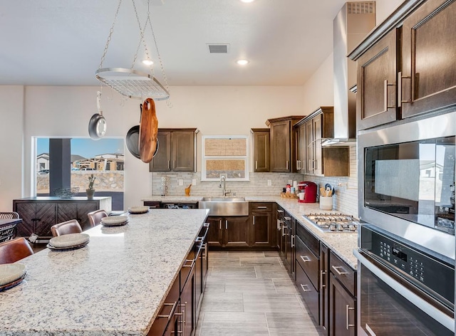 kitchen with appliances with stainless steel finishes, light stone counters, backsplash, wall chimney range hood, and sink