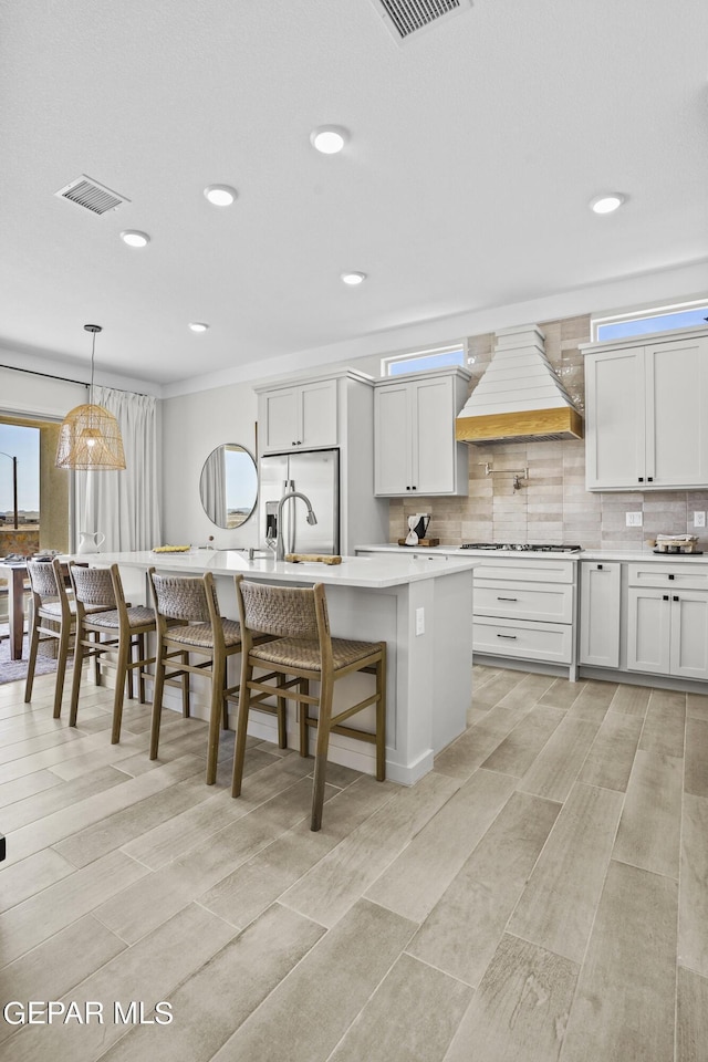 kitchen with white cabinets, decorative light fixtures, premium range hood, and a kitchen island with sink