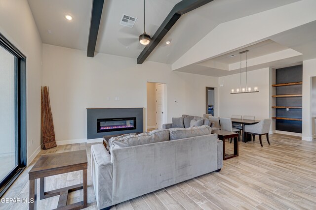 living area with visible vents, light wood-style flooring, a glass covered fireplace, ceiling fan, and vaulted ceiling with beams