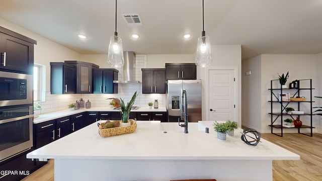 kitchen featuring wall chimney exhaust hood, appliances with stainless steel finishes, decorative light fixtures, and an island with sink