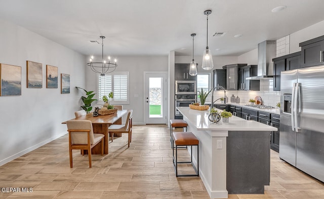 kitchen with appliances with stainless steel finishes, decorative light fixtures, a center island with sink, and wall chimney range hood