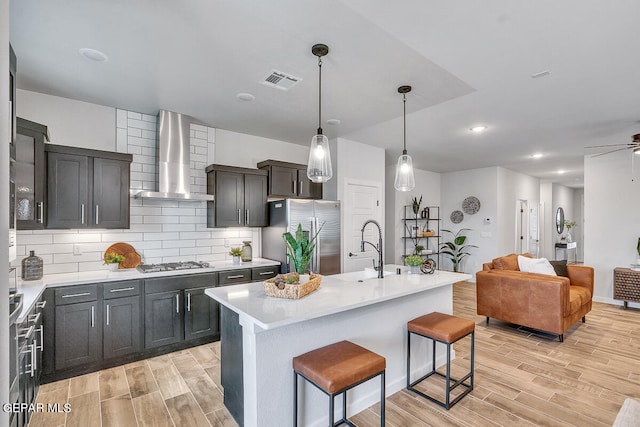 kitchen with stainless steel appliances, a kitchen bar, a center island with sink, and wall chimney range hood