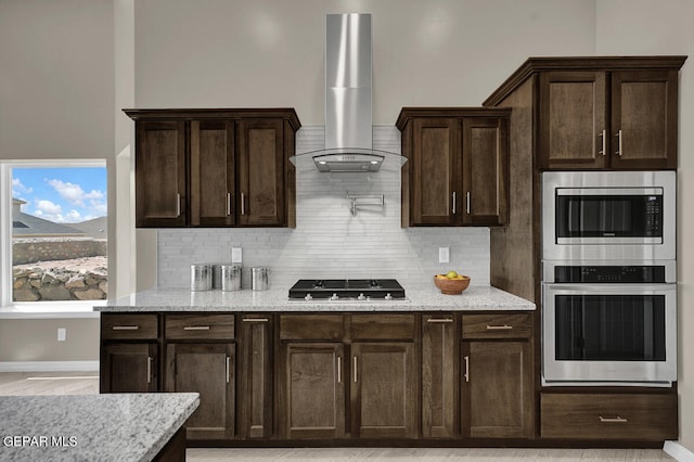 kitchen featuring dark brown cabinets, appliances with stainless steel finishes, light stone countertops, wall chimney range hood, and tasteful backsplash