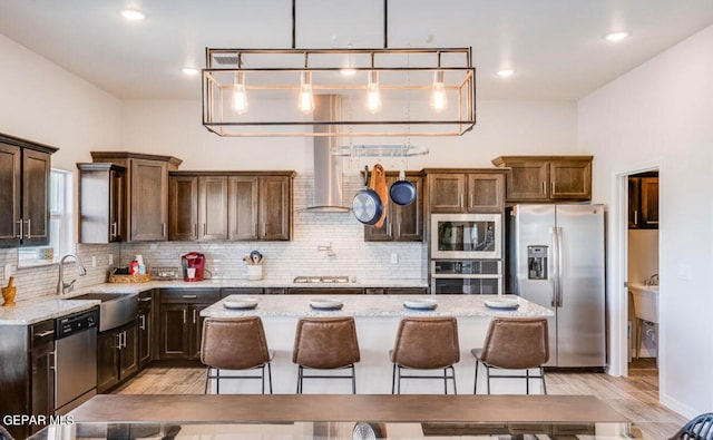 kitchen with stainless steel appliances, a kitchen island, wall chimney range hood, sink, and light stone counters