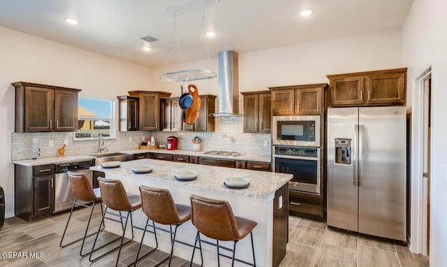 kitchen with stainless steel appliances, light stone counters, wall chimney range hood, a kitchen island, and a breakfast bar area